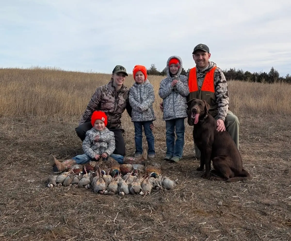 A family and their dog pose with harvested ring-necked pheasants and chukar partridges in a field.