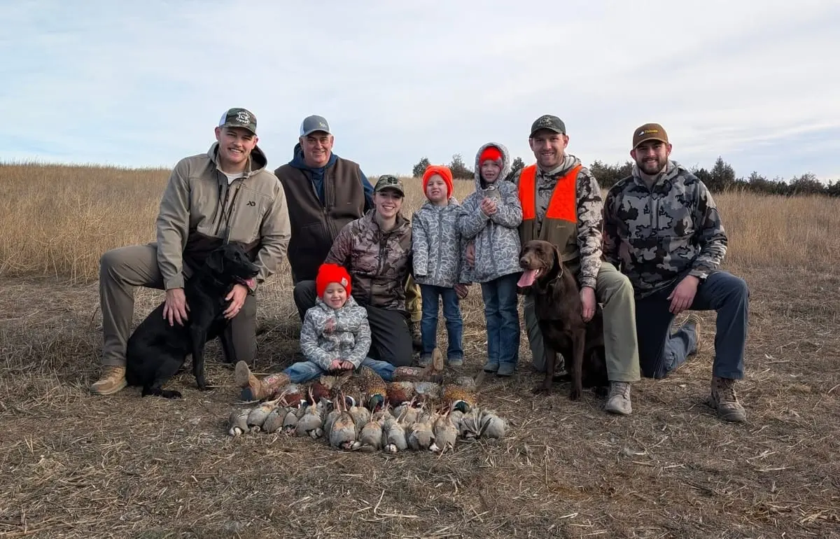 A group of hunters, including children and two dogs, pose with their harvest of ring-necked pheasants and chukar partridges in a field.