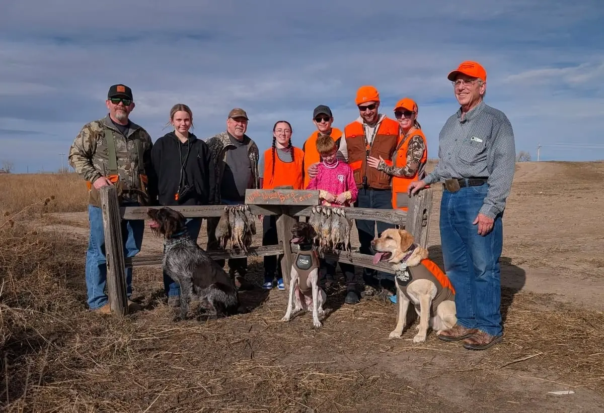 A group of hunters and two dogs pose with harvested chukar in a field.