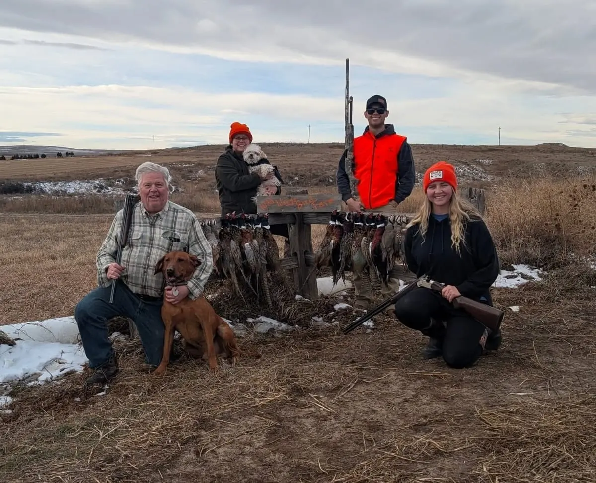 A group of four hunters and two dogs pose with harvested ring-necked pheasants on a cloudy day.
