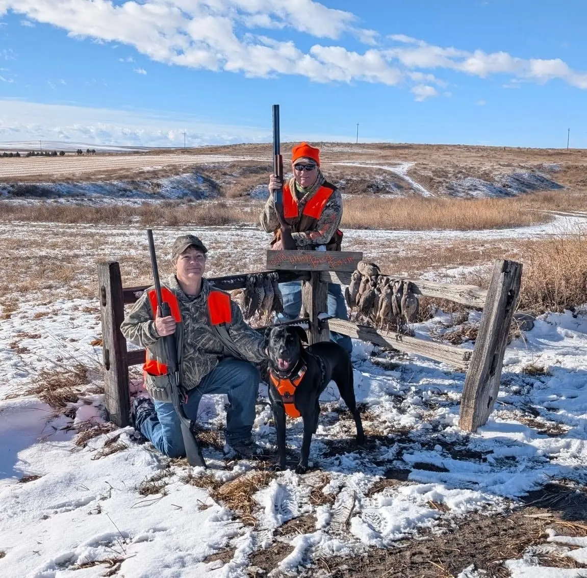 Two hunters and a dog pose with harvested chukar hanging on a fence in a snowy field.