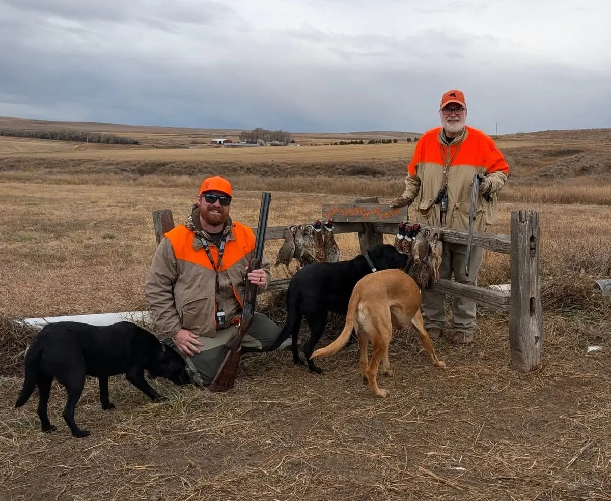 Two hunters pose with their dogs and harvested pheasants and chukar in a field.