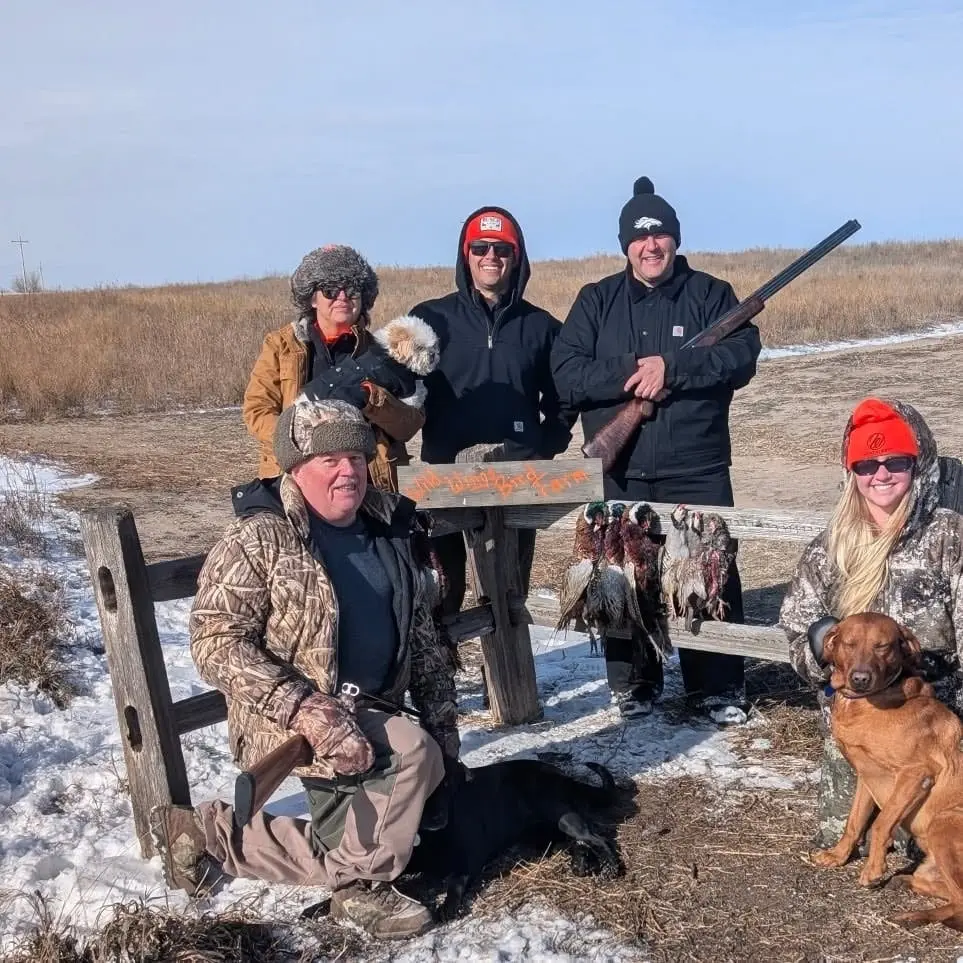 A group of hunters and their dogs pose with harvested Ring-necked Pheasants in a snowy field.