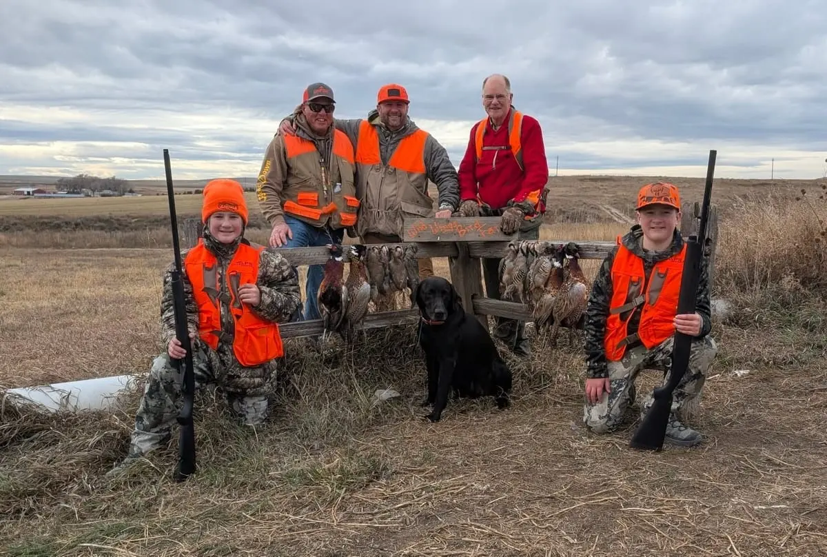 A group of hunters, including two young boys and a black dog, pose with harvested pheasants and chukar in a field.