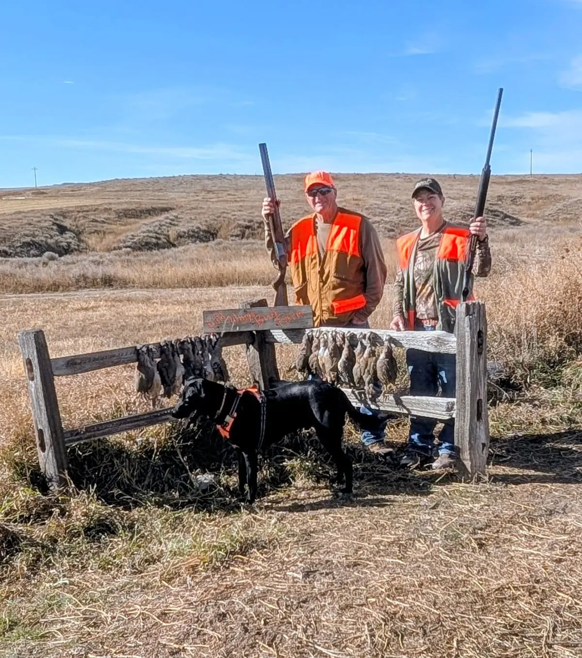 Two hunters and a black dog pose with harvested chukar partridges hanging on a wooden rack in a field.