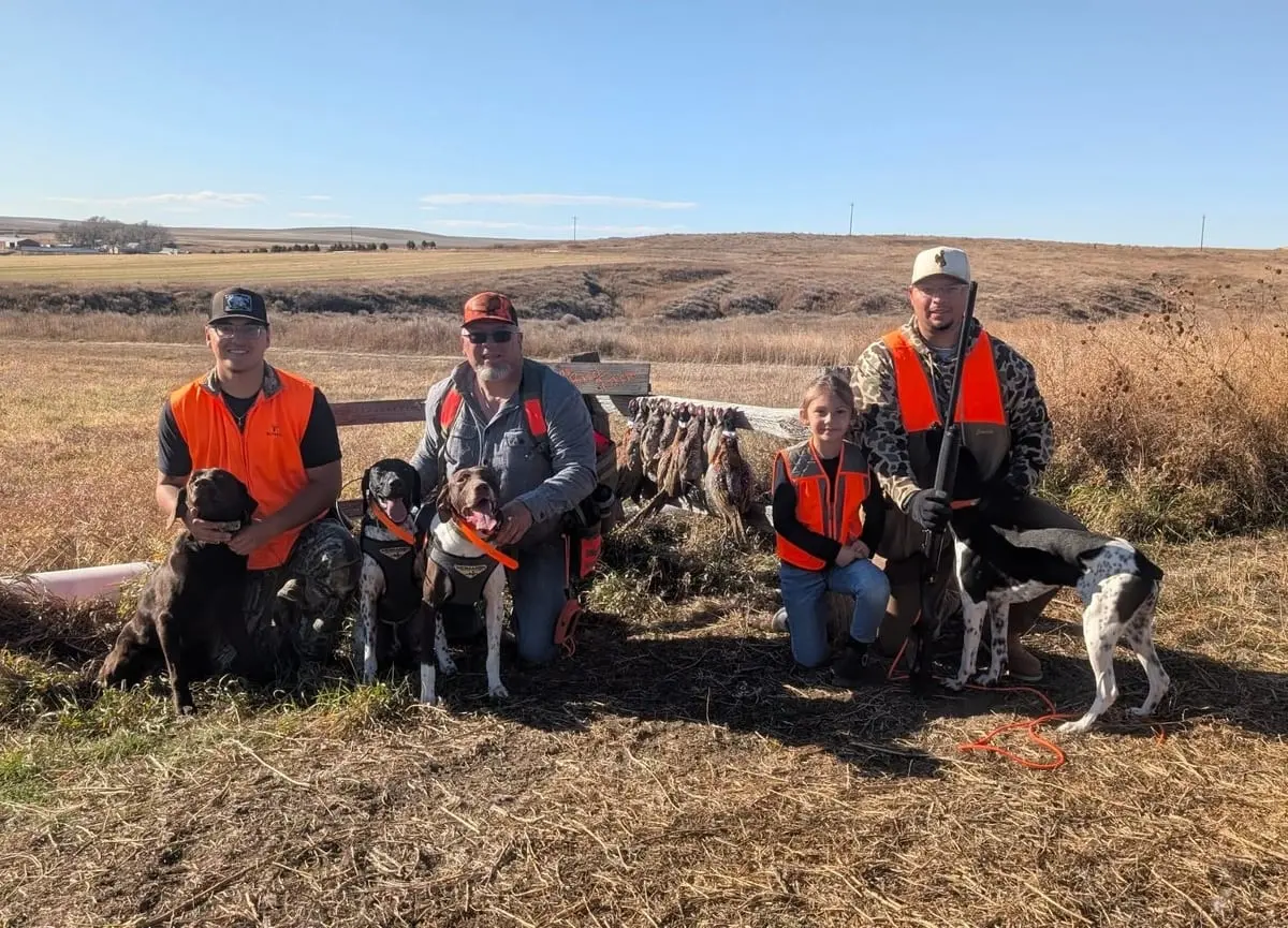 Three hunters and a child pose with their hunting dogs and harvested ring-necked pheasants in a field.