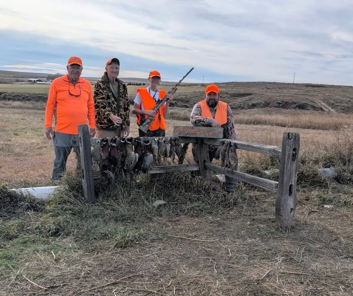 Four hunters pose with their harvested pheasants and chukar hanging on a wooden fence in a field.