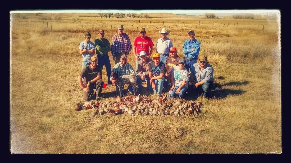 A group of hunters and a dog pose with their harvest of pheasants and chukar in a field.