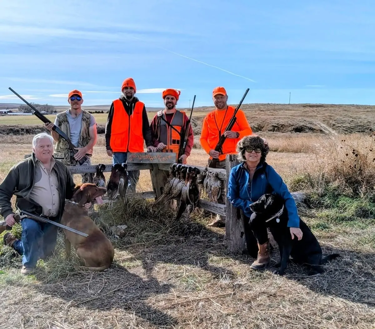 A group of hunters and their dogs pose with harvested pheasants and chukar in a field.