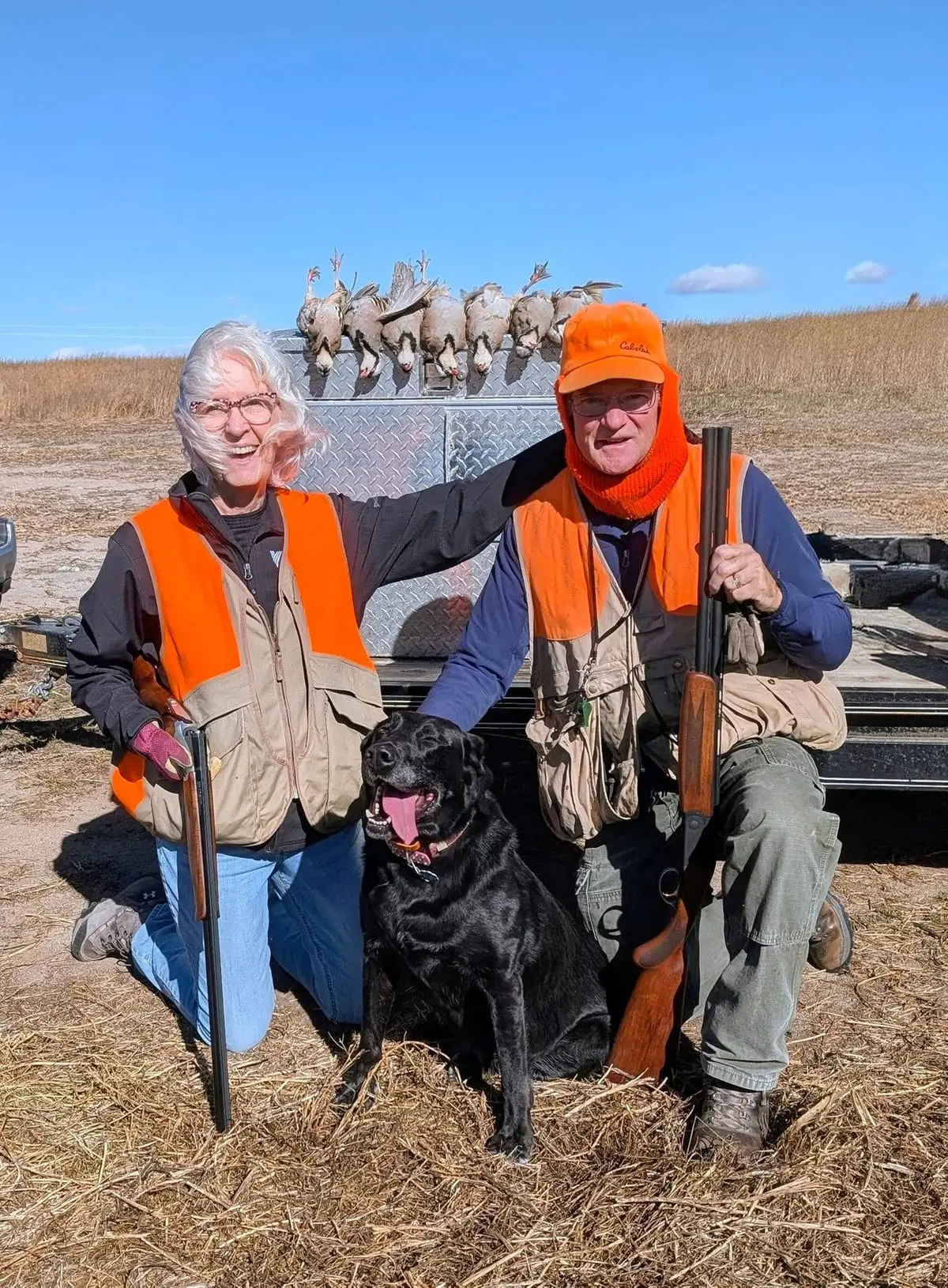 Two hunters and a black dog pose with several harvested chukar hanging from a trailer in a field.