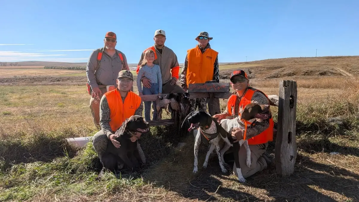 A group of hunters, including a child, pose with their hunting dogs and harvested ring-necked pheasants in a field.