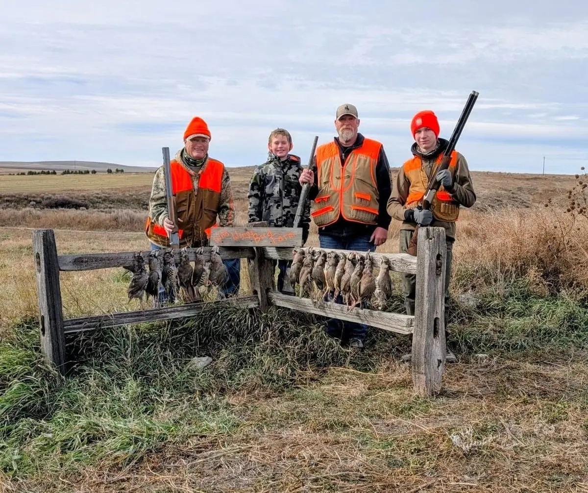 Four hunters pose with their harvested chukar partridges on a wooden rack in a field.