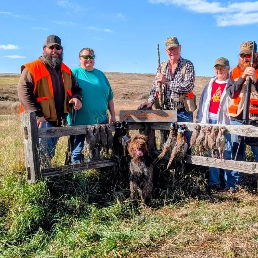 Hunters and a dog pose with harvested ring-necked pheasants and chukar partridges on a sunny day.