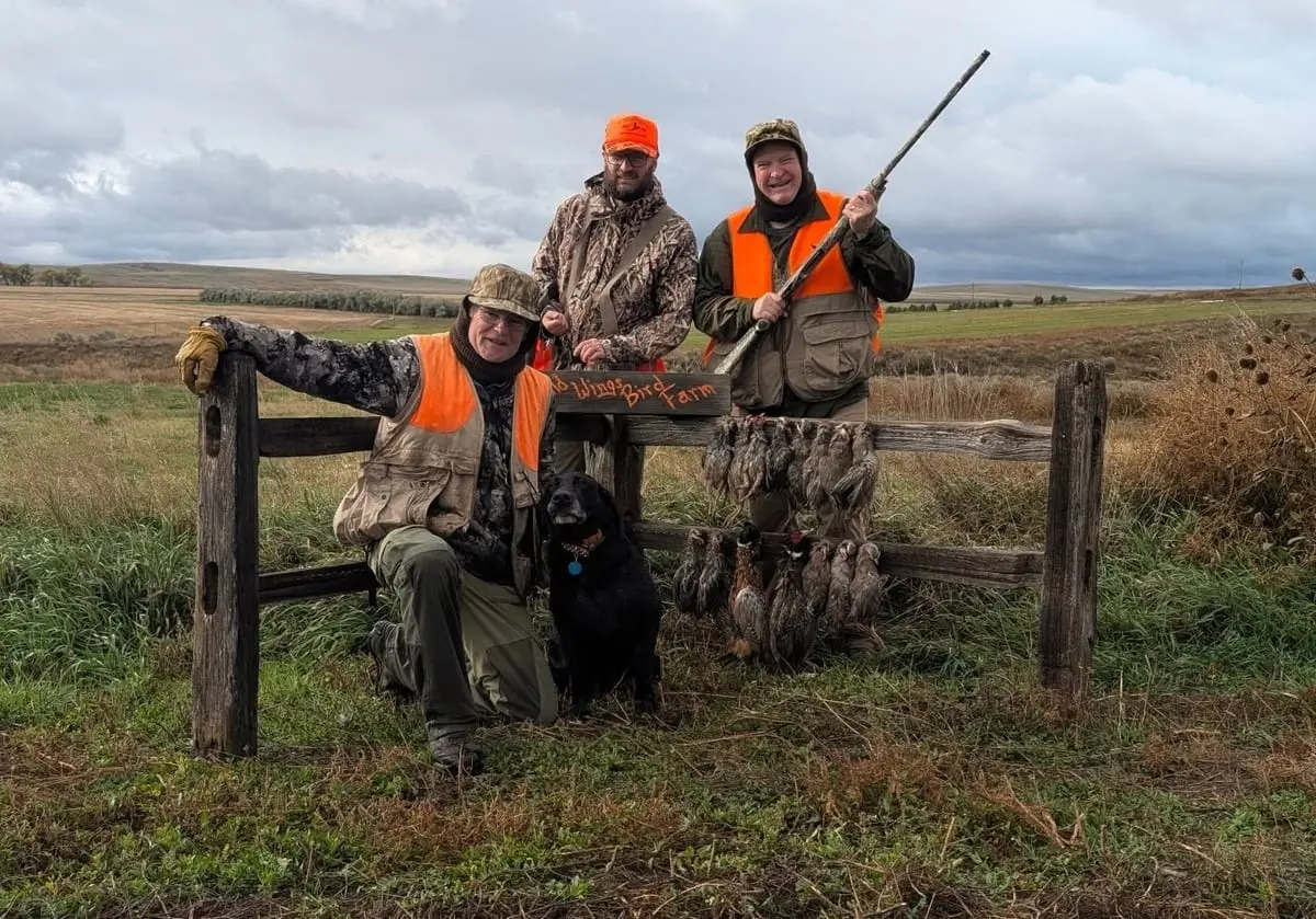 Three hunters and a dog pose with harvested chukar partridges hanging on a fence in a field.