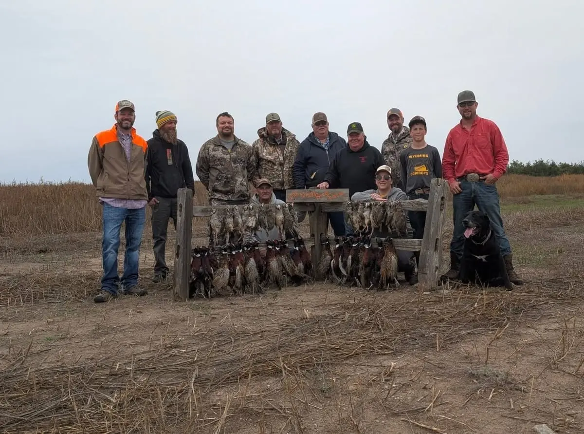 A group of hunters and a dog pose with their harvested pheasants and chukar in a field.