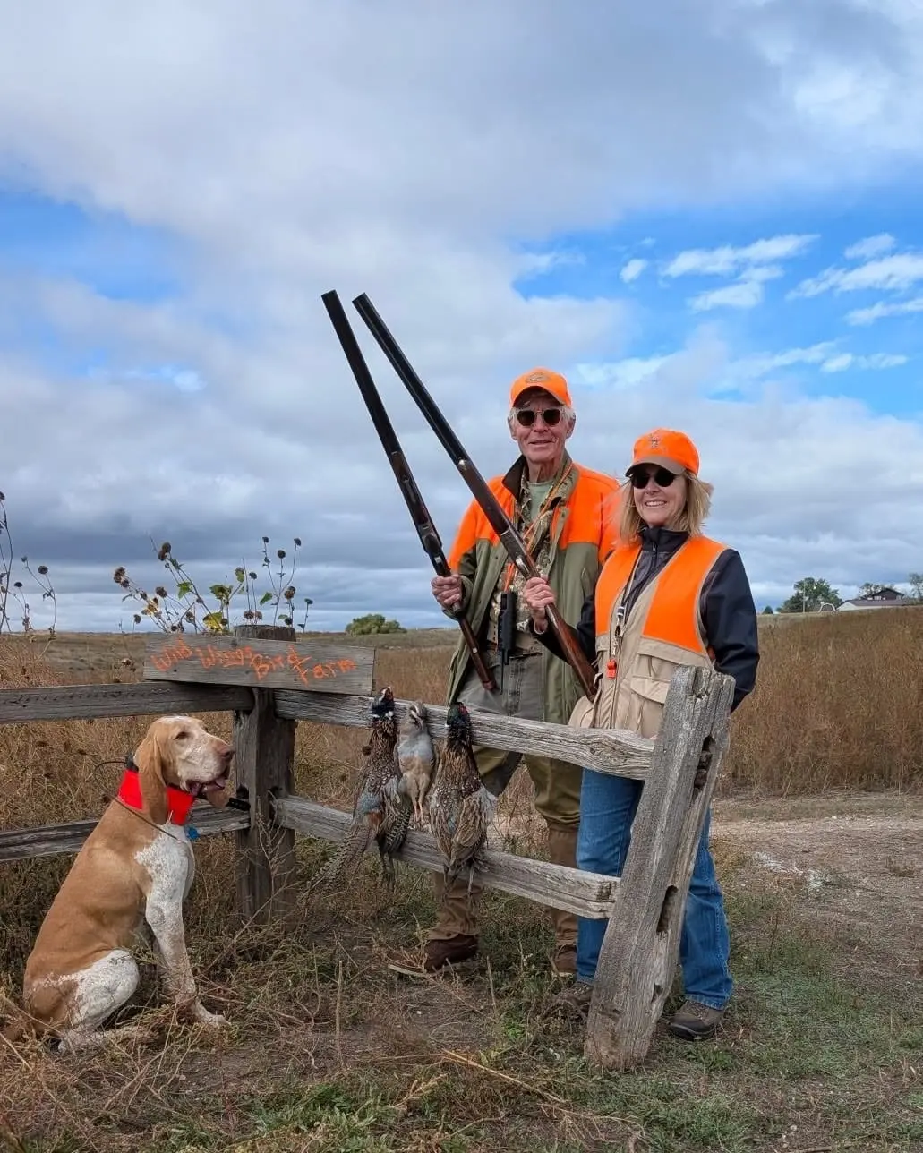 Two hunters and a dog pose with harvested pheasants and chukar hanging from a fence in a field.