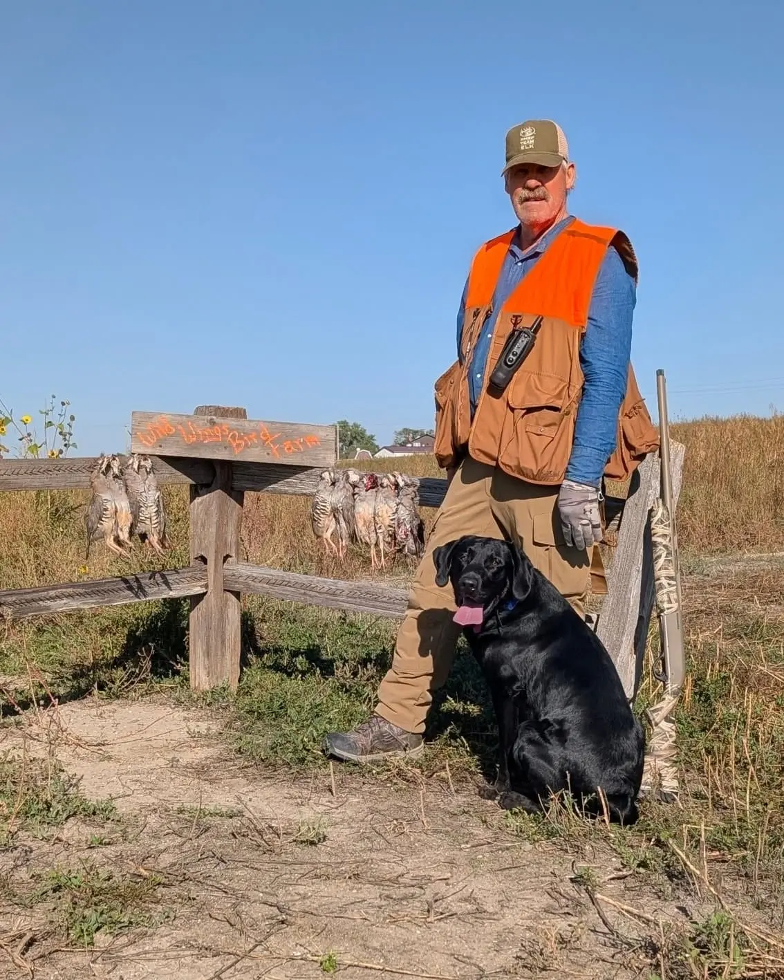 A man in hunting gear poses with his black dog and several harvested chukar hanging on a wooden fence in a field.