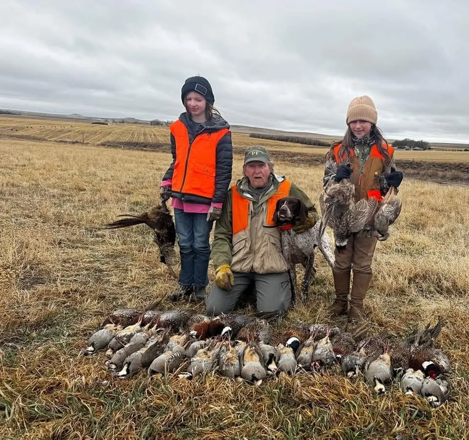 An adult male and two young girls pose with a hunting dog and a large harvest of ring-necked pheasants and chukar partridges in a field.