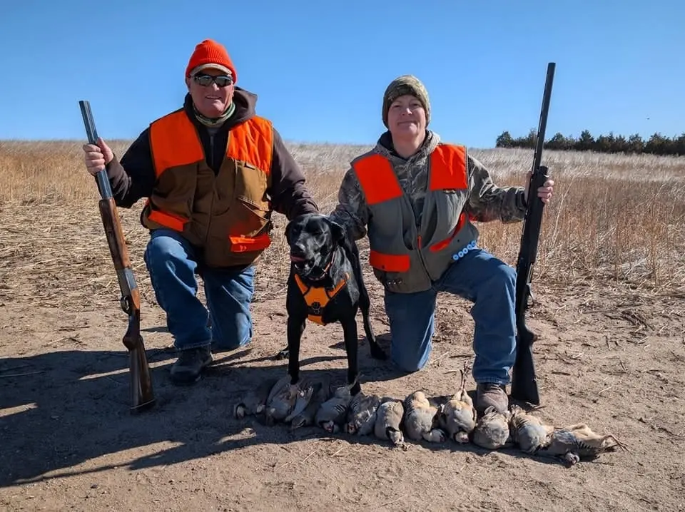 Two hunters and a dog pose with their harvested chukar partridges in a field.