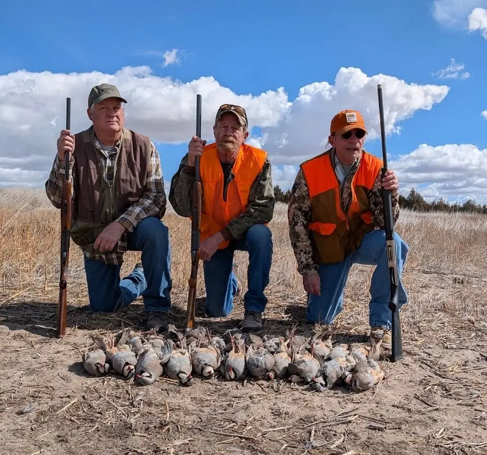 Three hunters kneel with their shotguns and a pile of harvested chukar partridges in a field under a blue sky.