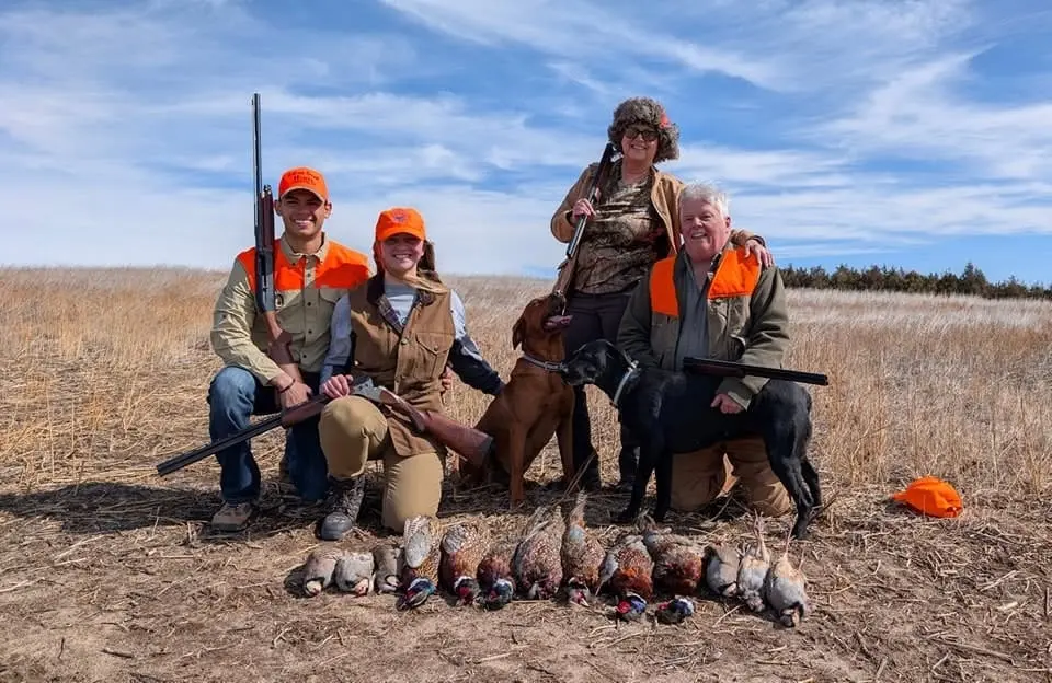 Four hunters and two dogs pose with their harvested pheasants and chukar in a field.