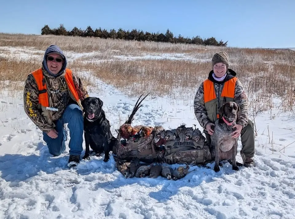 Two hunters and their dogs pose with a harvest of pheasants and chukar in a snowy field.