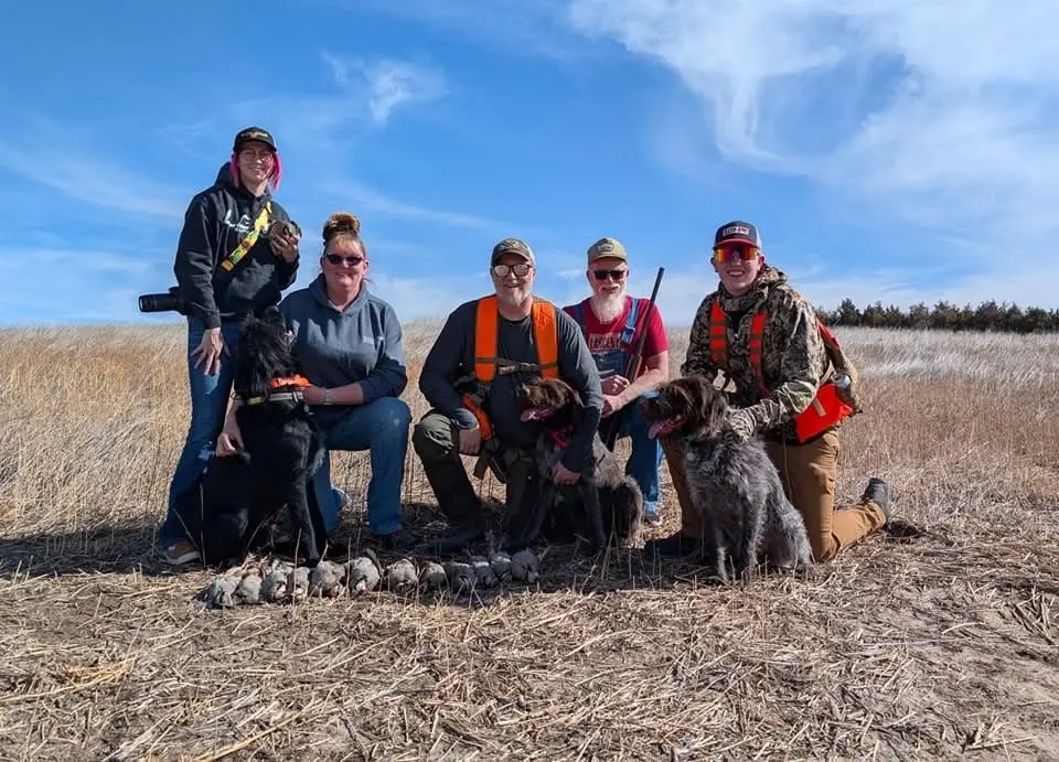 A group of five hunters and three dogs pose with harvested chukar in a field under a clear blue sky.
