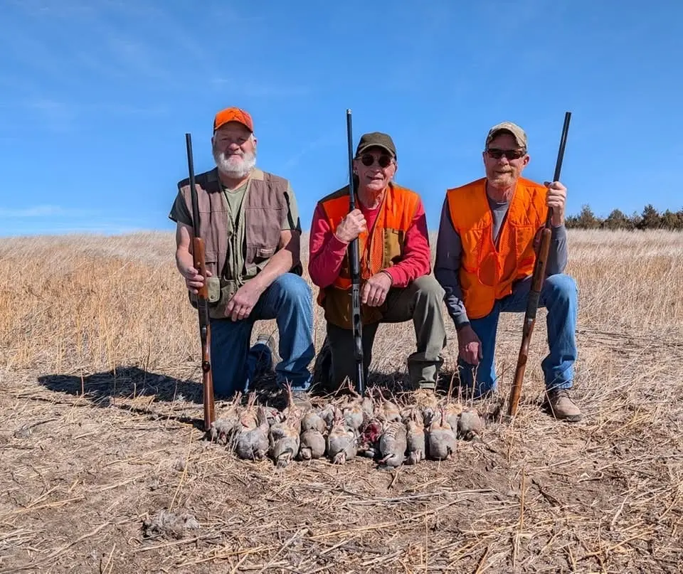 Three hunters kneel in a field with their shotguns and a pile of harvested chukar.