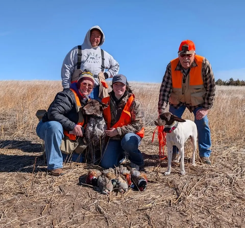 Four hunters and two dogs pose with harvested pheasants and chukar in a field.