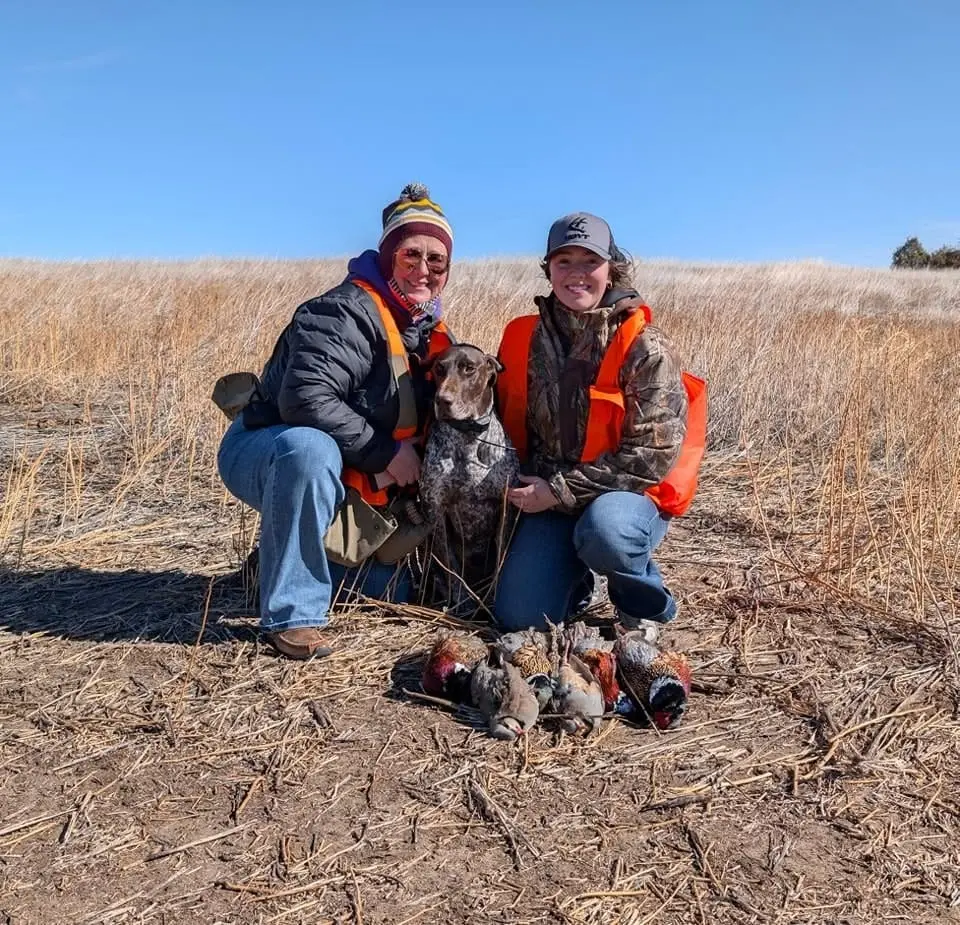 Two hunters and a dog pose with harvested ring-necked pheasants and chukar partridges in a field.