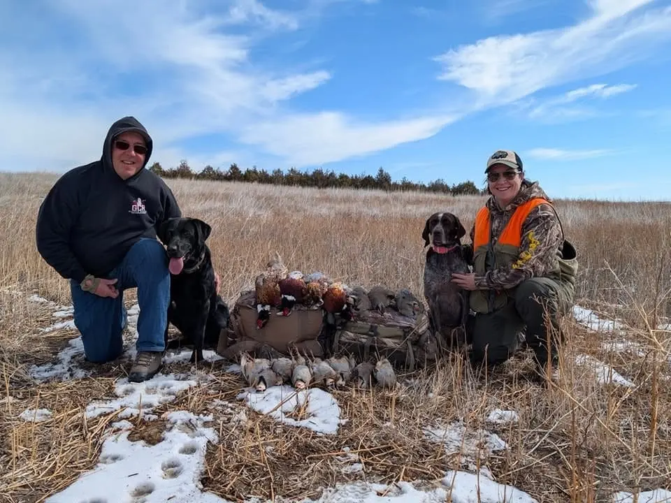 A man and a woman kneel in a snowy field with two hunting dogs and a pile of harvested ring-necked pheasants and chukar partridges.