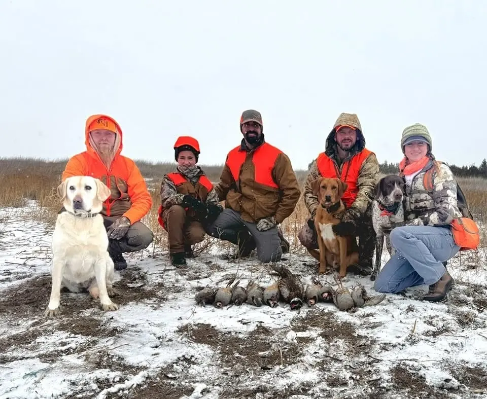 A group of hunters and their dogs pose with harvested chukar and a pheasant in a snowy field.