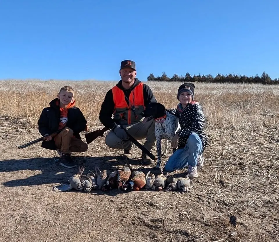 A man, two children, and a dog pose with harvested ring-necked pheasants and chukar partridges in a field.