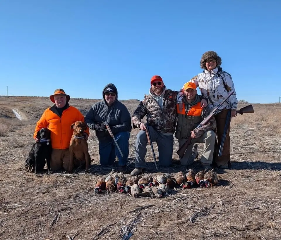 A group of five hunters and two dogs pose with their harvested pheasants and chukar in a field.