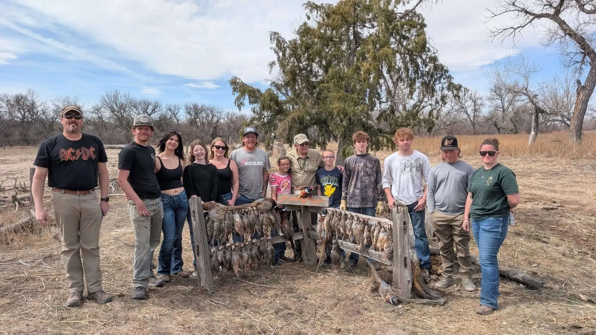 A group of people pose with harvested pheasants and chukar on a rack in a field.