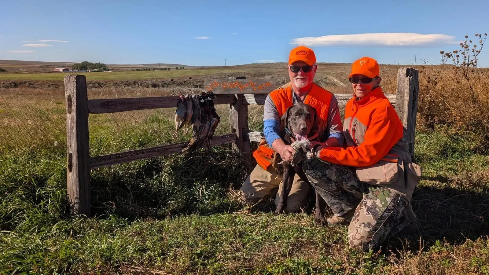 Two hunters and a dog pose with harvested pheasants hanging on a wooden fence in a field.