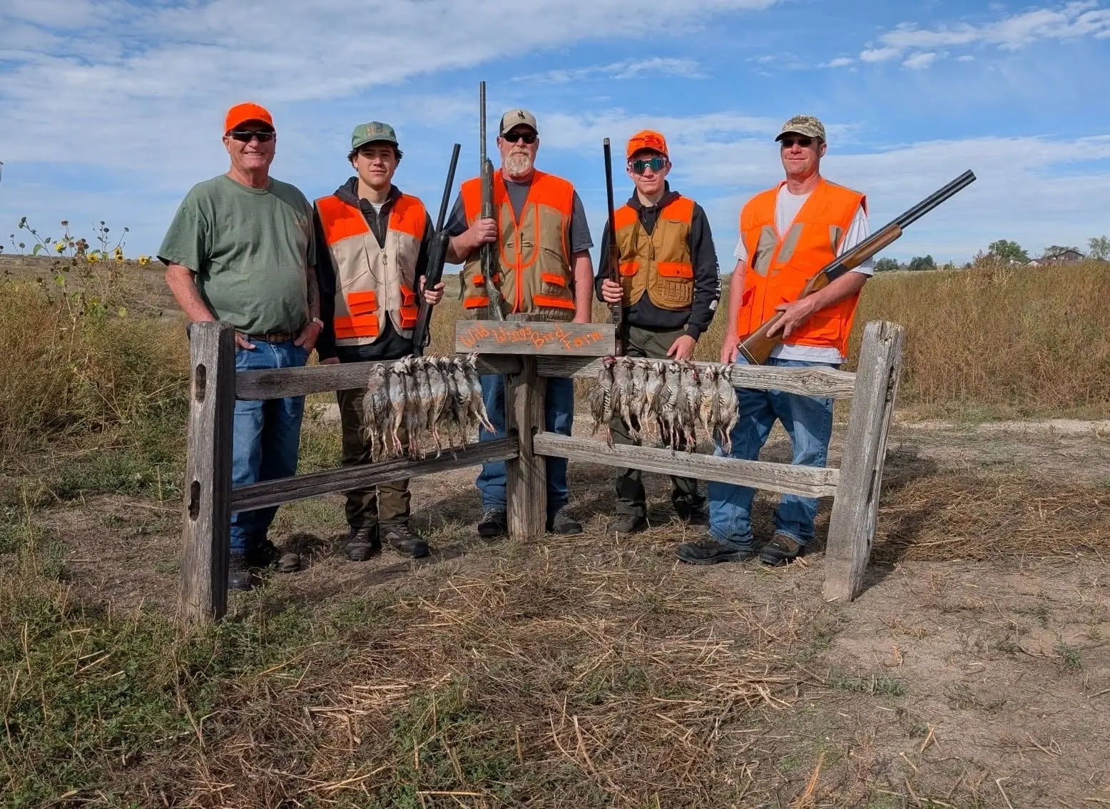Five hunters pose with their harvested chukar partridges hanging on a wooden fence.