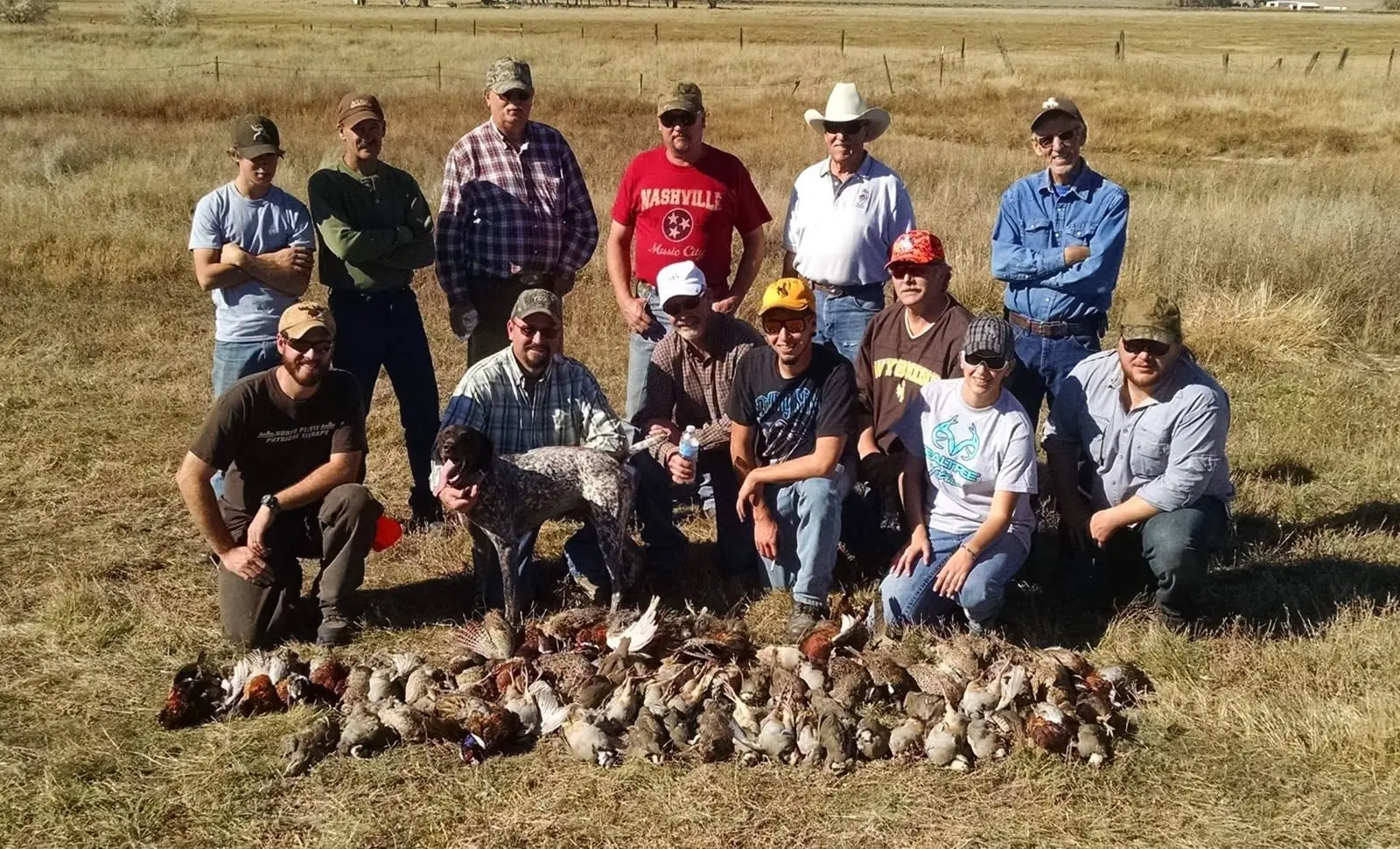 A large group of hunters, including a dog, pose in a field with their harvest of ring-necked pheasants and chukar partridges.